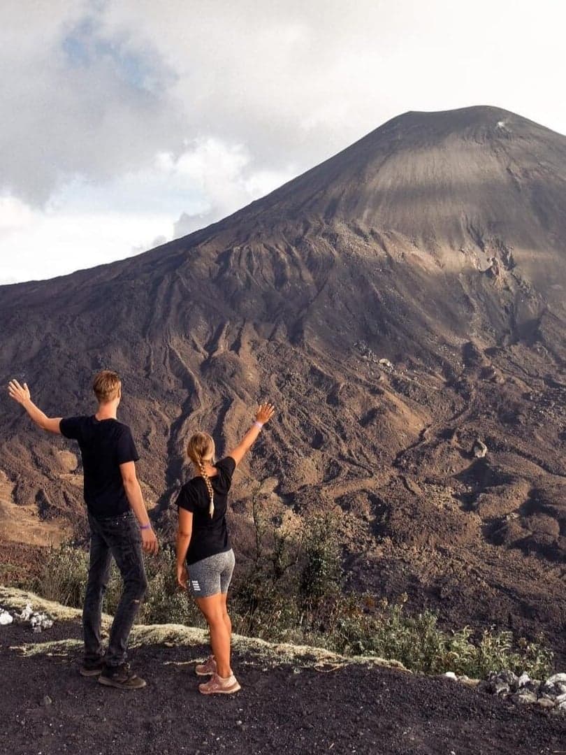 Two hikers raising their arms in front of the dark volcanic cone of Pacaya