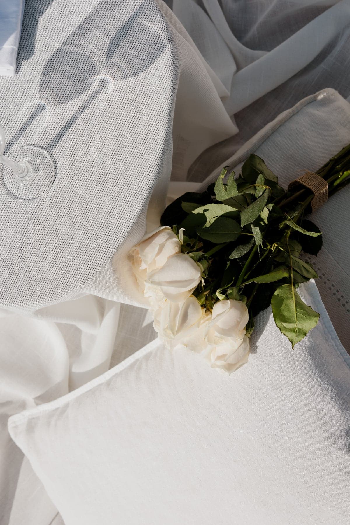Bouquet of white roses resting on white linen beside the shadow of a wine glass